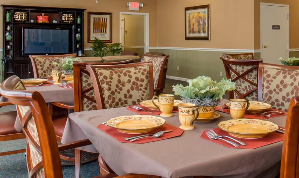 A dining area in a senior living facility with tables set for four people. Each table has a brown tablecloth, red placemats, yellow plates, matching mugs, and silverware. There is a decorative green plant centerpiece on the table. In the background, there is a dark wooden entertainment center with a TV and decorative items, beige walls with framed artwork, and a door labeled 'Restroom'.