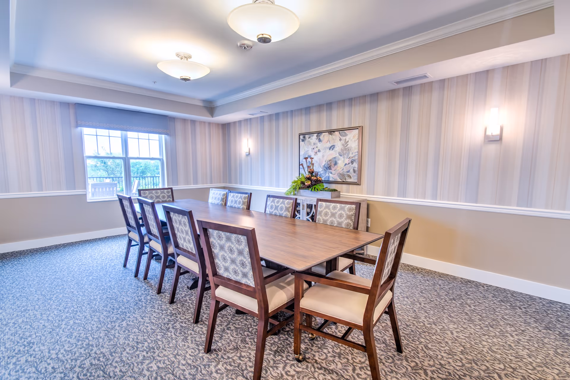 Bright dining room with a long wooden table and upholstered chairs, patterned carpet, striped wallpaper, and a window.