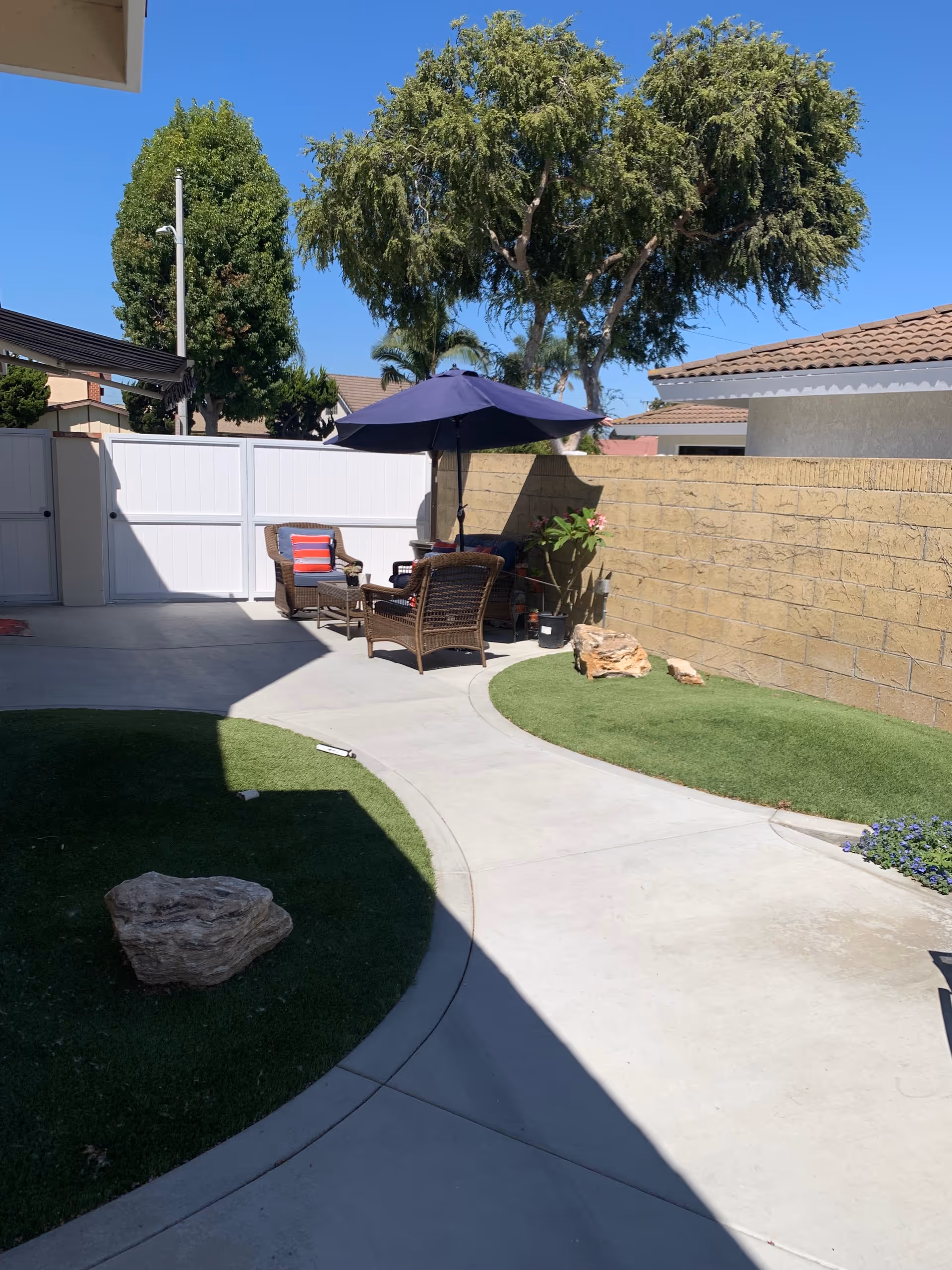 Outdoor patio area with a curved concrete walkway, artificial grass patches, a few large rocks, and a seating area with wicker chairs and a table under a blue umbrella. Trees and a white fence are visible in the background under a clear blue sky.