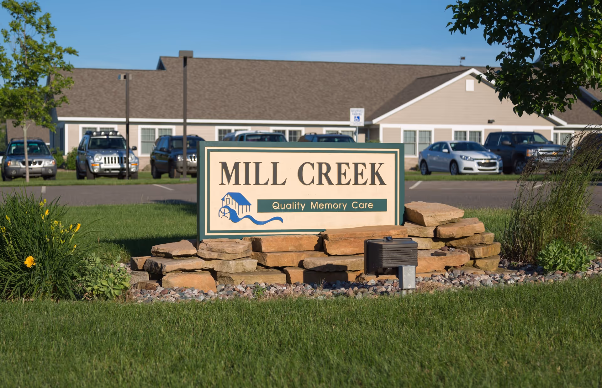 A sign for Mill Creek Quality Memory Care is displayed on a stone base in front of a senior living community building with parked cars and green grass around.