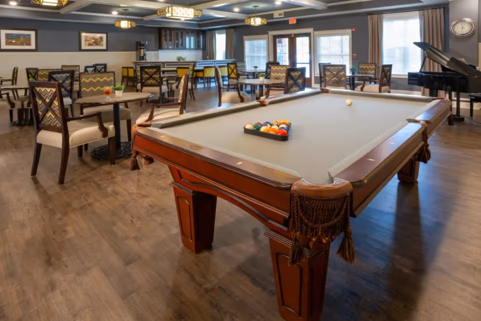 A spacious common area featuring a pool table with a full rack of billiard balls, surrounded by multiple tables and chairs with patterned upholstery. The room has wooden flooring, large windows with curtains, a grand piano in the corner, and soft ceiling lighting fixtures.