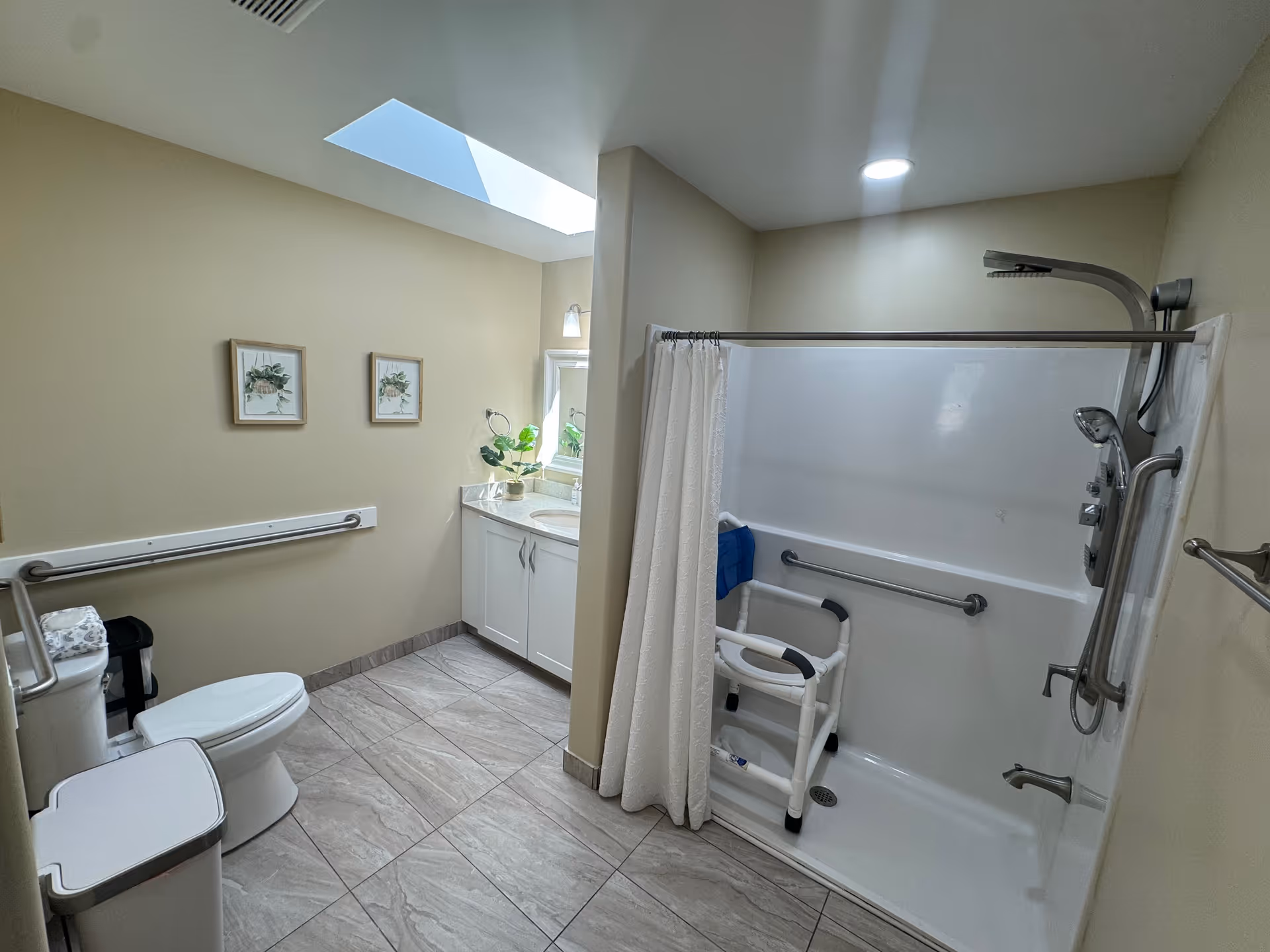 A spacious, accessible bathroom with beige walls and tiled floor. The room features a white toilet with grab bars on the wall, a white vanity with a sink and a mirror above it, and a walk-in shower with a white curtain, grab bars, a shower chair, and a handheld showerhead. There are two framed botanical prints on the wall and a skylight providing natural light.