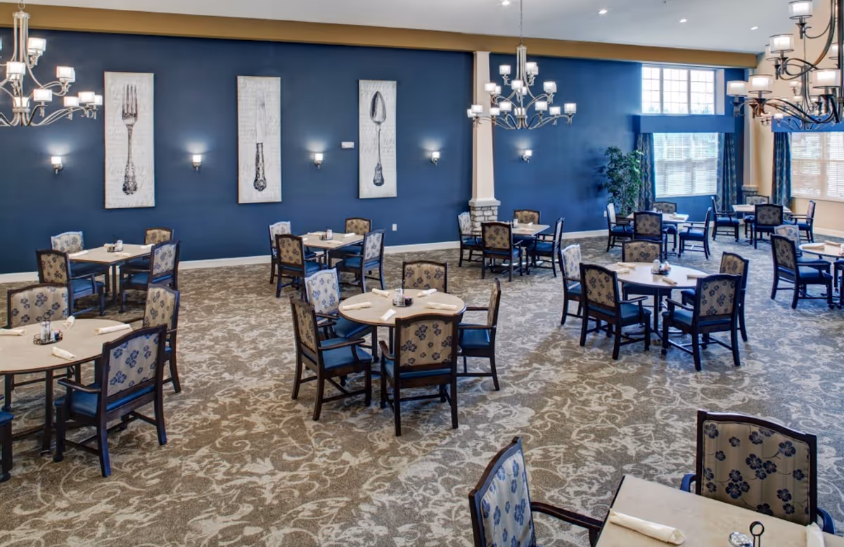 A spacious senior living dining room with multiple round and square tables, each set with napkins and condiments. The room features patterned carpet, blue walls adorned with large artwork of a fork, knife, and spoon, and elegant chandeliers hanging from the ceiling. Large windows allow natural light to fill the space.