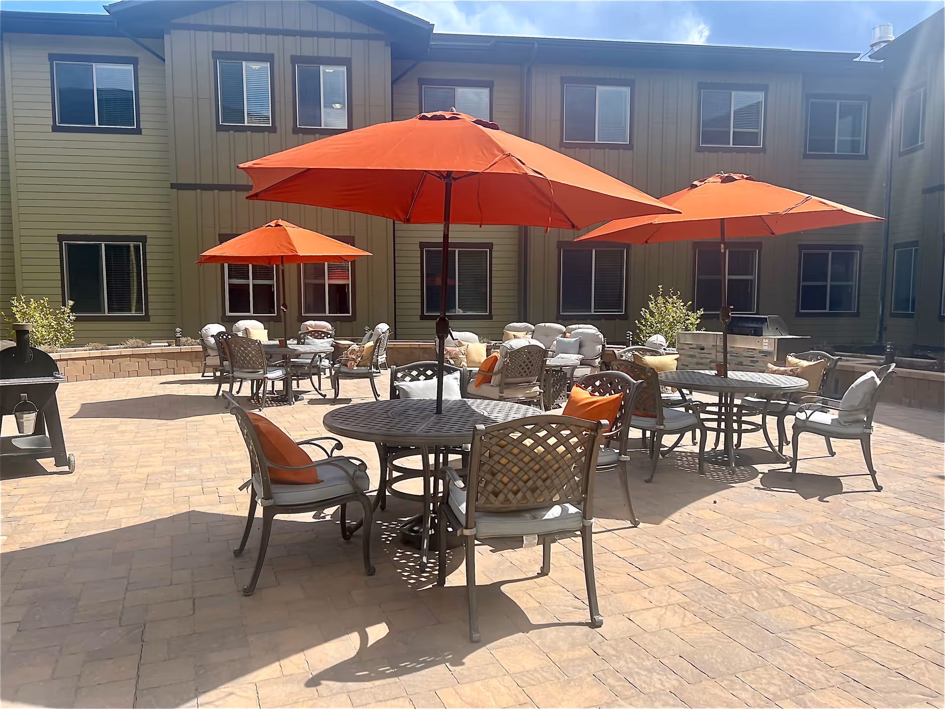 Outdoor patio area at Spring Gardens Senior Living Draper with several round metal tables and chairs, each table shaded by large orange umbrellas. Cushions and pillows are placed on the chairs, and there is a barbecue grill and a pizza oven on the paved patio. The building with multiple windows surrounds the patio.