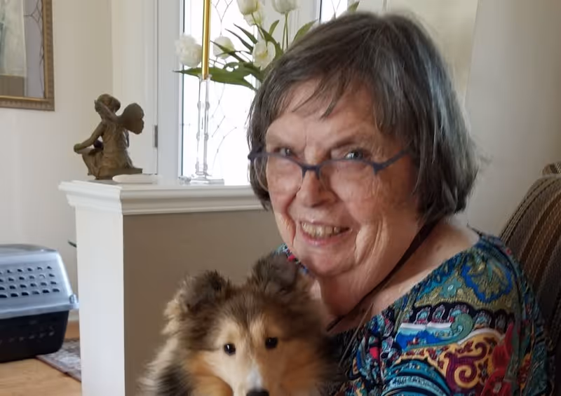 An elderly woman with glasses and short gray hair smiling while holding a fluffy dog in a cozy living room setting. Behind her is a white half-wall with a small statue and a vase with flowers, and a pet carrier is visible on the floor.