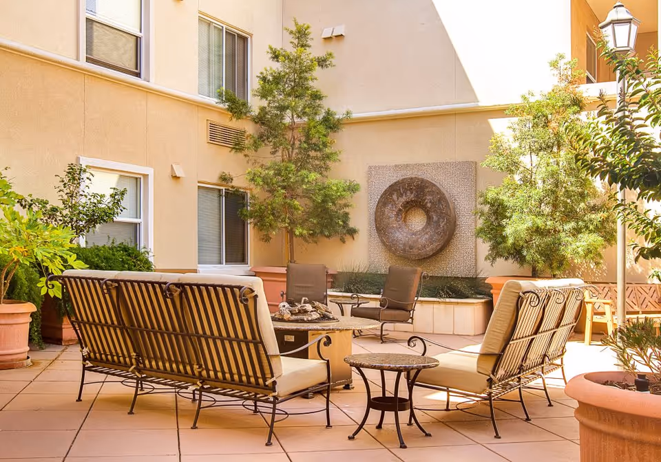 Outdoor courtyard area with cushioned metal benches and chairs arranged around a fire pit table. The space is surrounded by potted plants and trees, with a decorative circular wall fountain mounted on the beige building wall. A lamp post is visible on the right side.