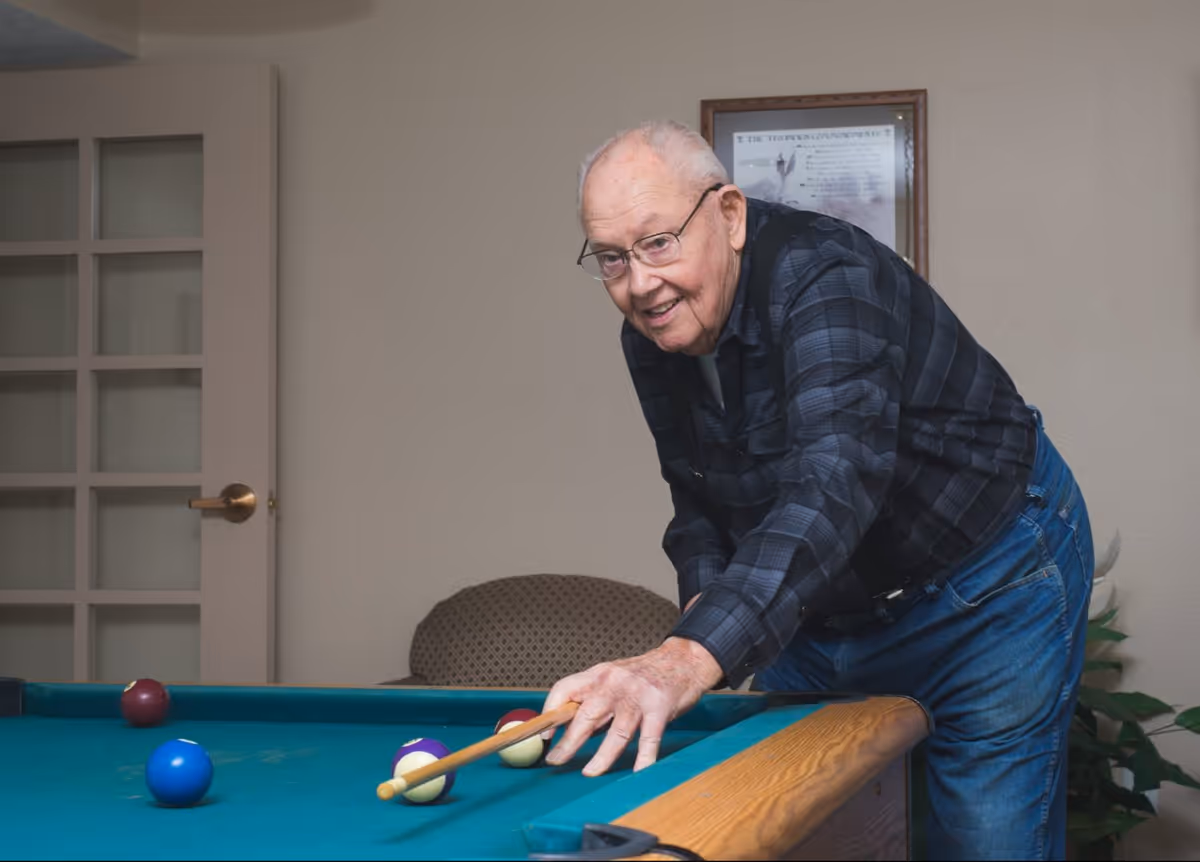 An elderly man wearing glasses, a dark plaid shirt, and blue jeans is playing pool indoors. He is leaning over a pool table, aiming with a cue stick at the balls on the table. Behind him is a beige wall with a framed picture and a door with glass panels.