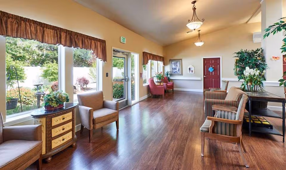 A bright and spacious common area in an assisted living facility with large windows overlooking a garden. The room features wooden flooring, several comfortable chairs arranged along the walls, a small decorative cabinet, and potted plants. There are two hanging light fixtures on the ceiling and a red door at the far end of the room.