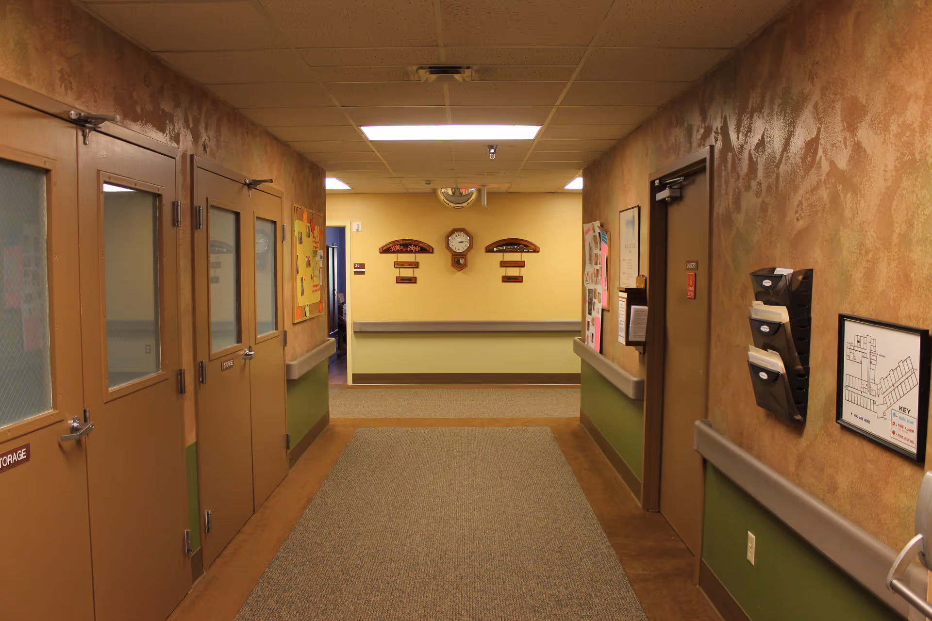 A hallway in a healthcare facility with beige and green walls, several closed doors on both sides, bulletin boards, a wall clock, and a floor plan mounted on the wall. The hallway is carpeted with a patterned runner and has overhead fluorescent lighting.