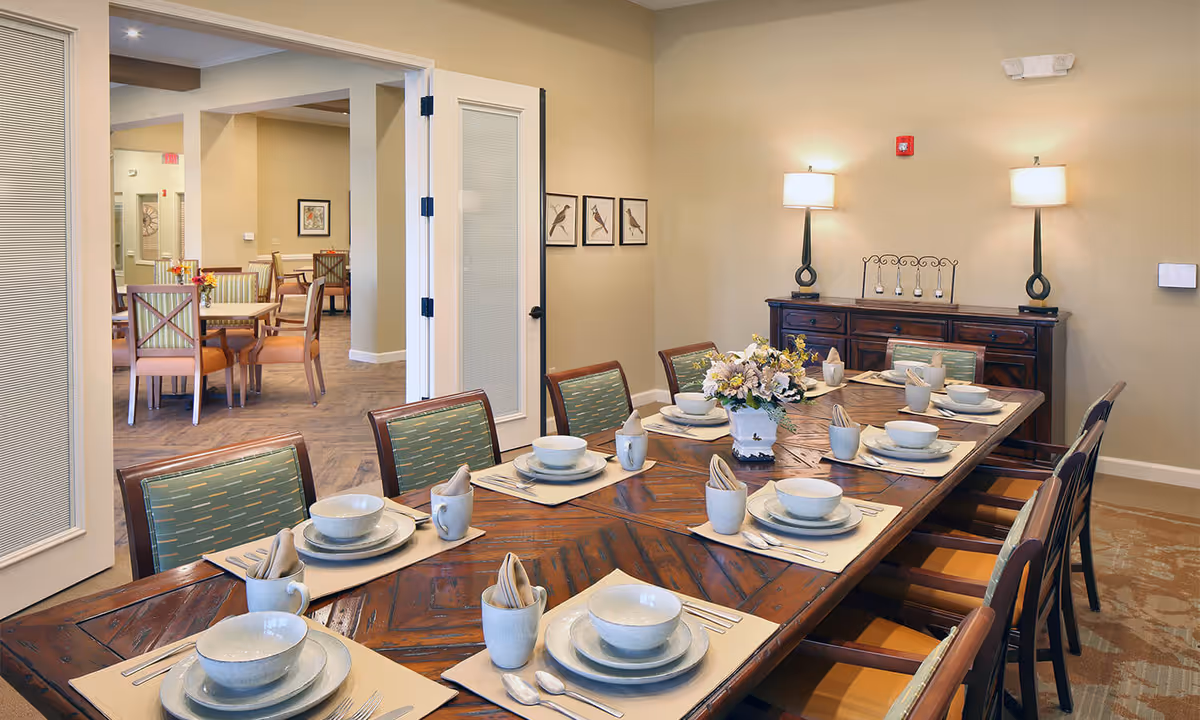 A well-lit dining room with a long wooden table set for eight people, each place setting including a bowl, plate, mug, napkin, and cutlery. The chairs have green patterned cushions. In the background, there is a sideboard with two lamps and decorative items, and an open doorway leading to another dining area with additional tables and chairs.