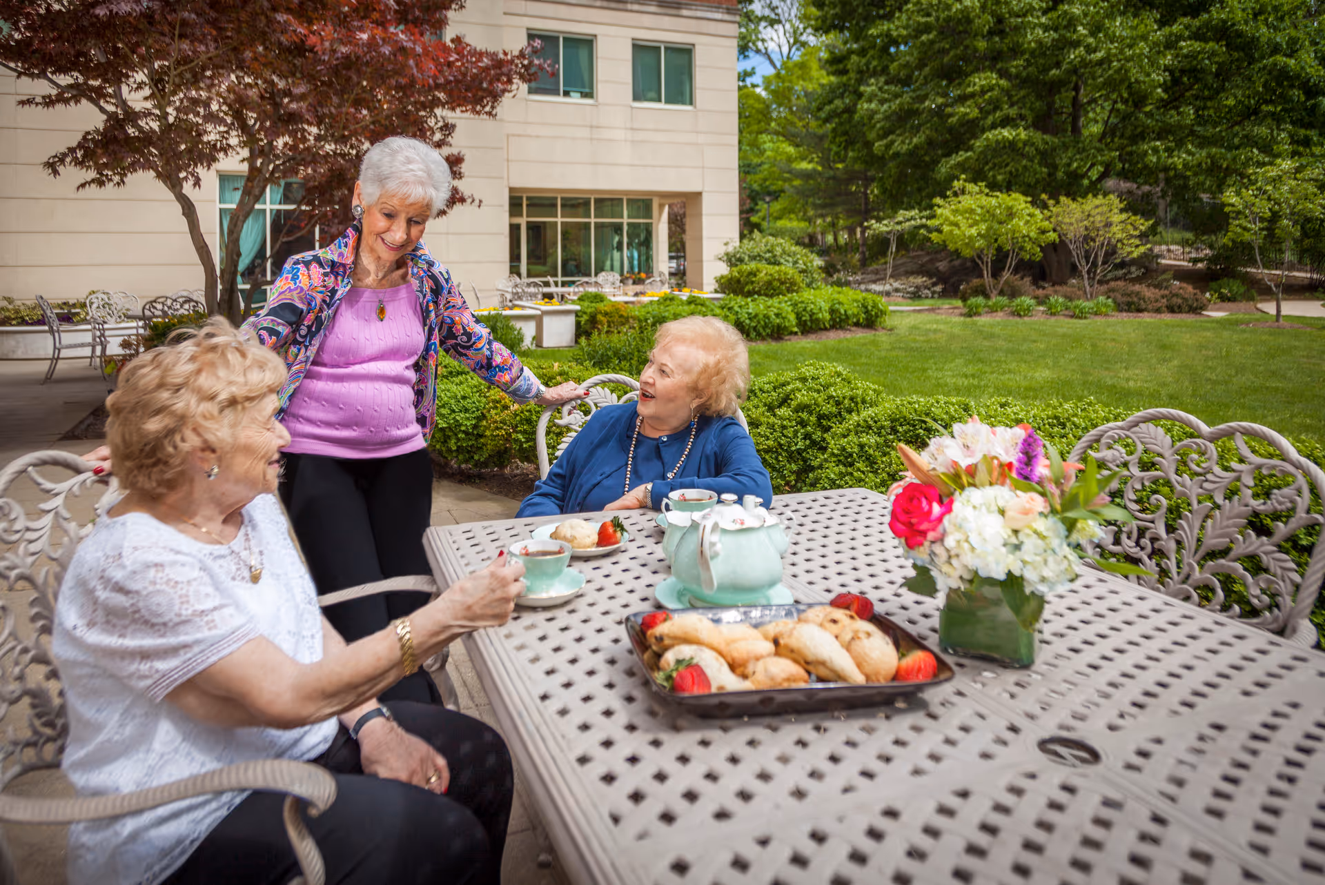 Three elderly women enjoying tea and pastries at an outdoor metal table in a garden area with green bushes and trees. One woman is standing and smiling while the other two are seated and engaged in conversation. A vase with colorful flowers is on the table along with a tray of pastries and a teapot set.