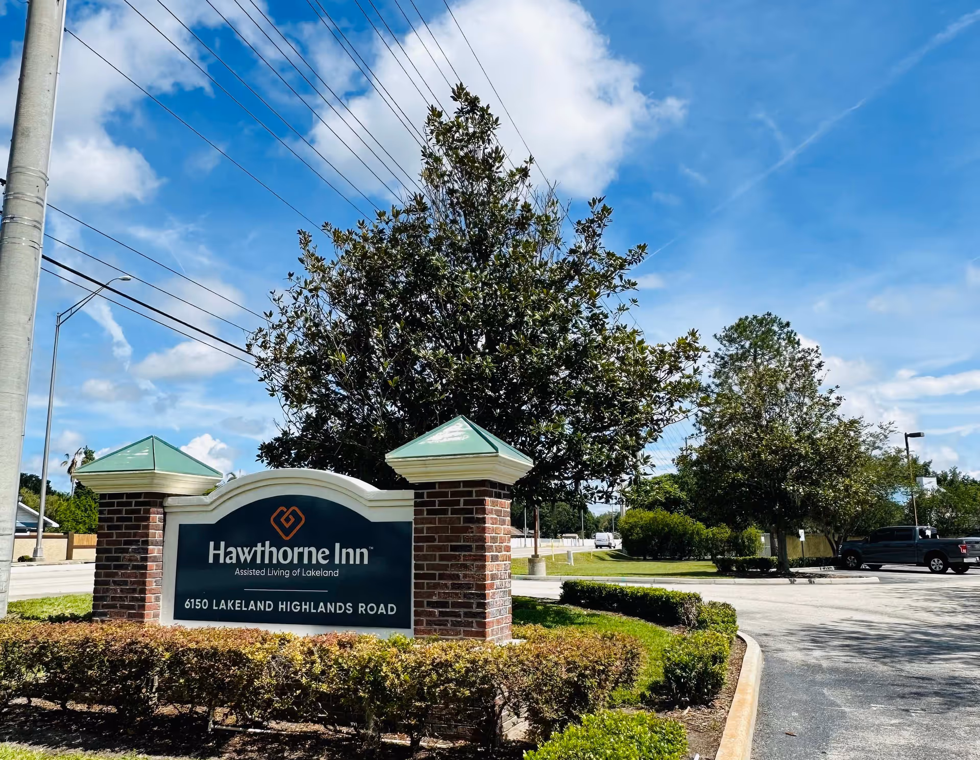 Brick entrance sign reading 'Hawthorne Inn Assisted Living of Lakeland' with landscaping, trees, a parking area and a blue sky overhead.