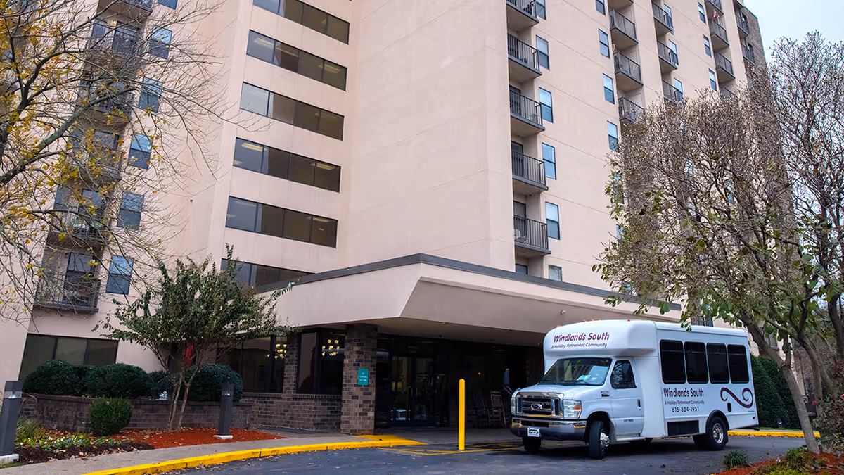 Exterior view of a multi-story senior living facility building with a covered entrance. A white shuttle bus labeled 'Windlands South A Holiday Retirement Community' is parked near the entrance. Trees and landscaping surround the driveway.