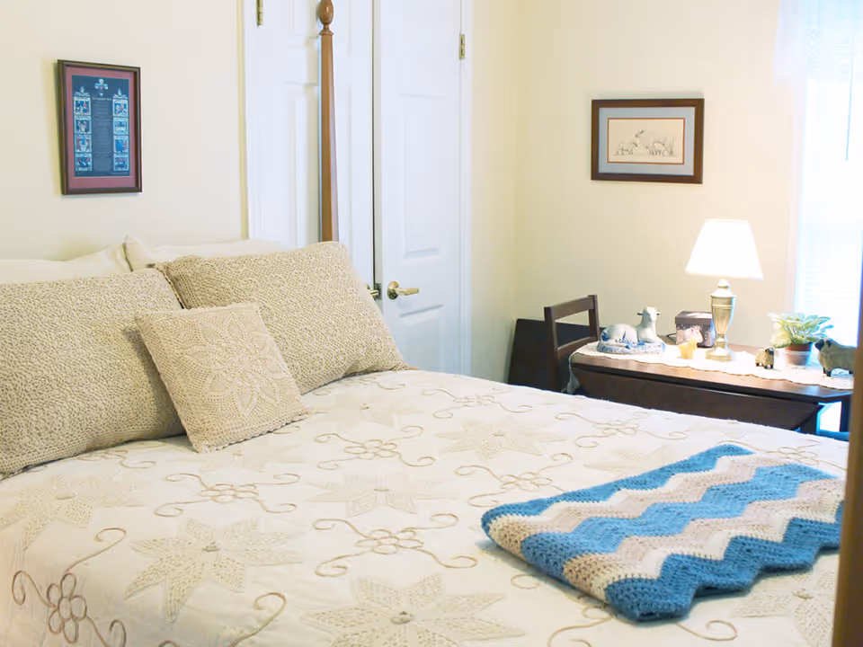 A cozy bedroom with a neatly made bed featuring beige patterned pillows and a quilt with floral designs. A blue and white crocheted blanket is folded at the foot of the bed. In the background, there is a wooden chair and a table with a lamp, decorative items, and a small plant. Two framed pictures hang on the light-colored walls.