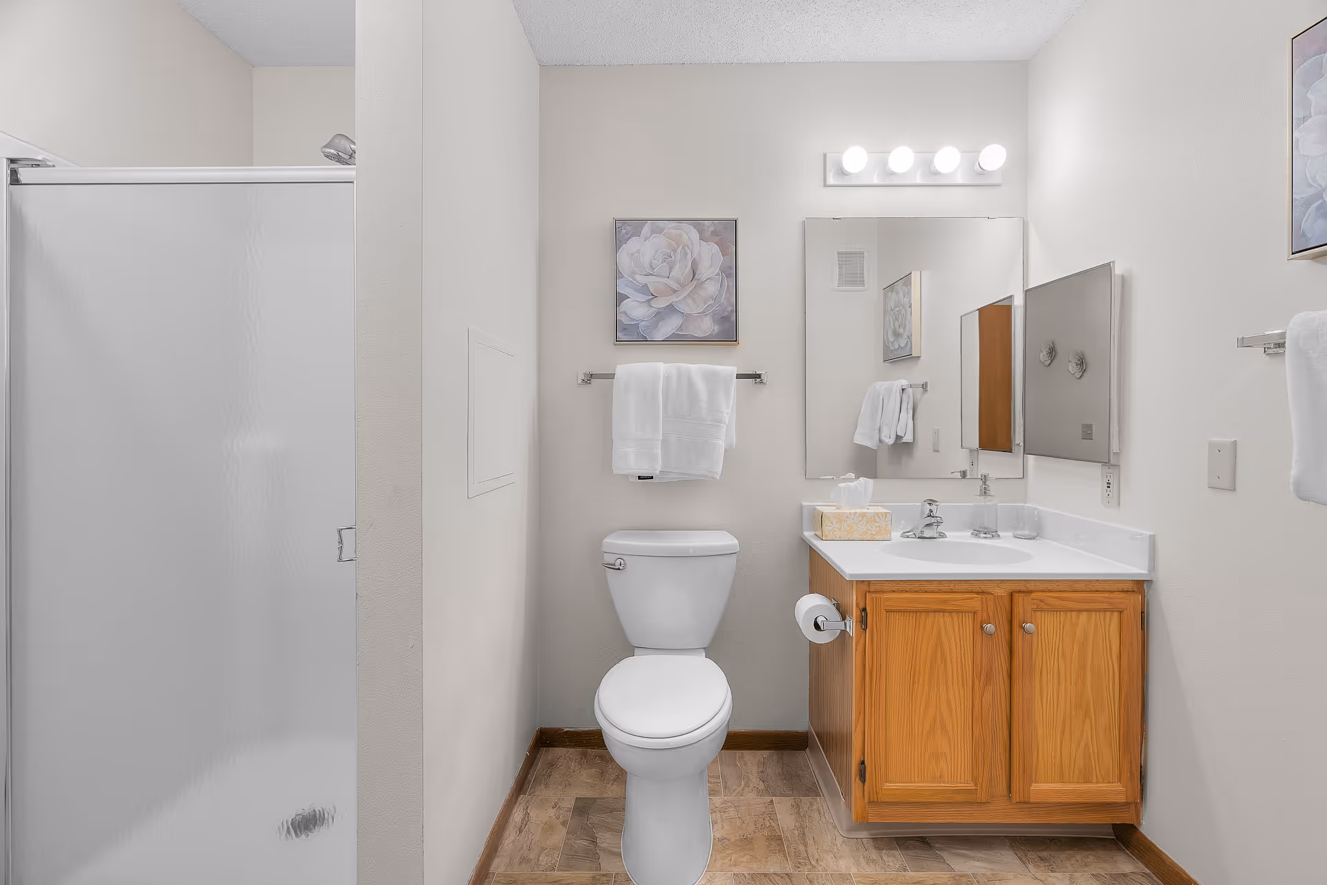 A neat bathroom showing a toilet centered, wooden vanity with sink and mirror to the right, towel and artwork above, and a frosted-glass shower stall to the left.