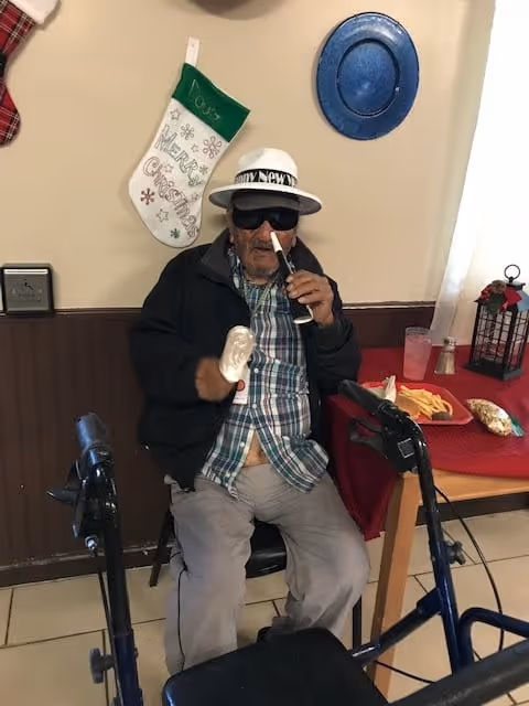 An elderly man wearing a white hat, dark sunglasses, a plaid shirt, and a black jacket is sitting on a chair next to a walker. He is holding a white object in one hand and a small black and white object near his nose with the other hand. Behind him on the wall is a green and white Christmas stocking and a blue decorative plate. To his right is a table with a red tablecloth, a plate of fries, a drink, and a lantern decoration.