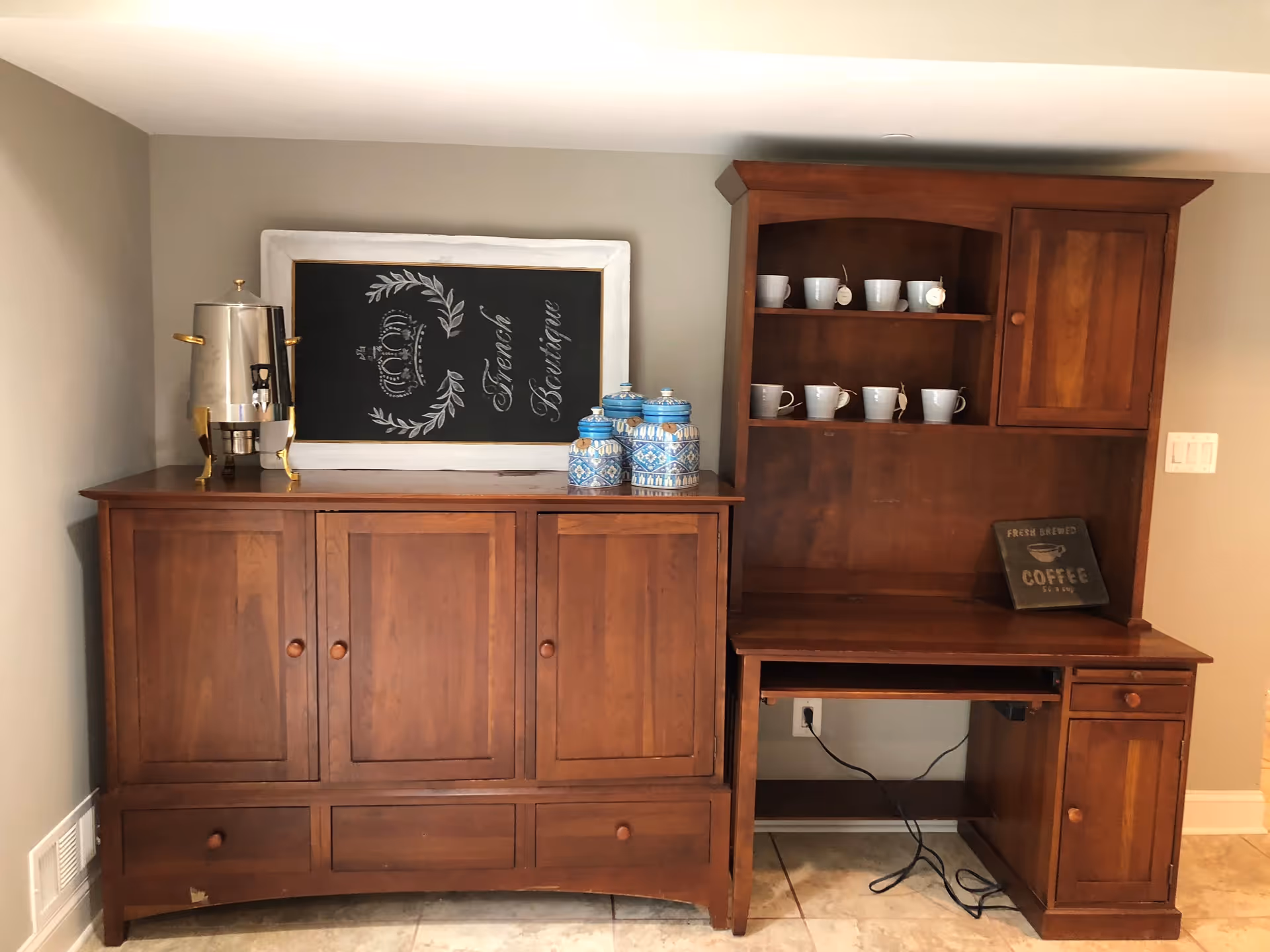 A wooden cabinet and hutch set against a beige wall. The cabinet has a silver beverage dispenser and three blue and white decorative jars on top. Above the cabinet is a framed chalkboard with the words 'La French Boutique' and a crown design. The hutch has shelves with white mugs and a small sign that reads 'Fresh Brewed Coffee'. The floor is tiled and there is an electrical outlet with a black cord plugged in beneath the hutch.