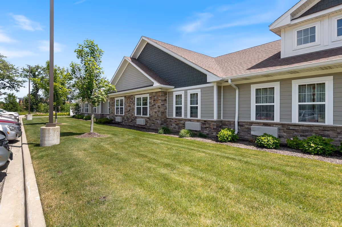 Exterior view of a single-story building with gray siding and stone accents, multiple white-framed windows, a well-maintained lawn, small shrubs, and a parking area with cars on the left side under a clear blue sky.
