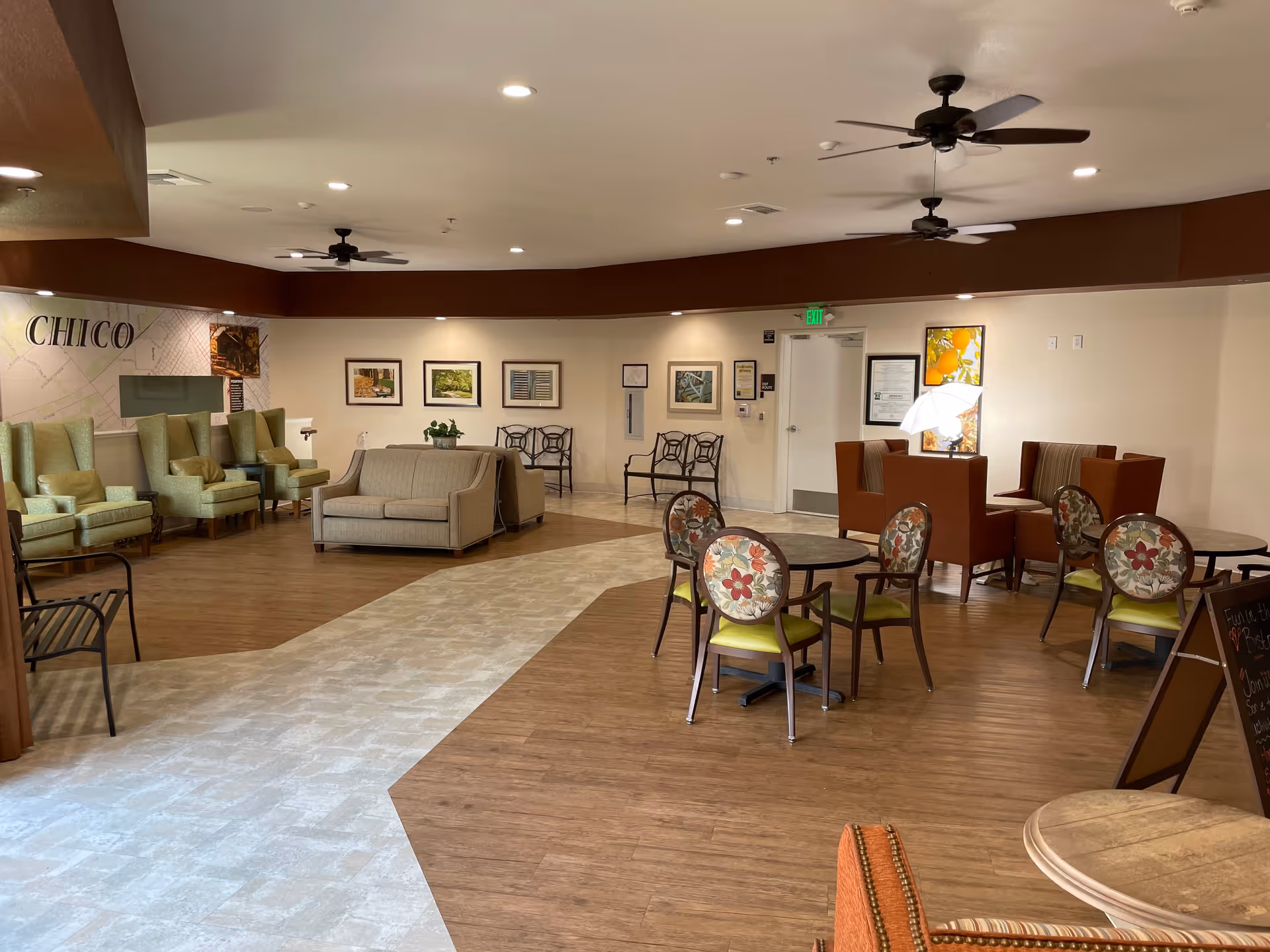 A spacious senior living facility common area with a mix of seating options including armchairs, sofas, and round tables with floral cushioned chairs. The floor has a combination of wood and tile patterns. The walls are decorated with framed pictures and a large map labeled 'CHICO'. Ceiling fans and recessed lighting illuminate the room.