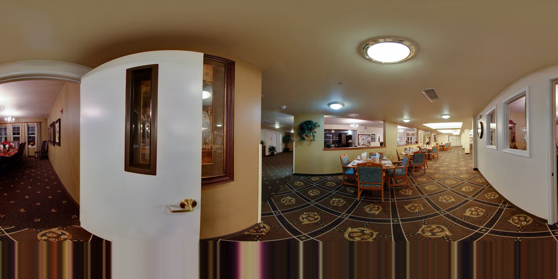 Panoramic view of a senior living facility dining area with multiple tables and chairs arranged neatly. The carpet has a patterned design, and the walls are painted in a light color. There is a door partially open leading to another room with a dining table and chairs. Ceiling lights illuminate the space, and some framed artwork and plants decorate the area.