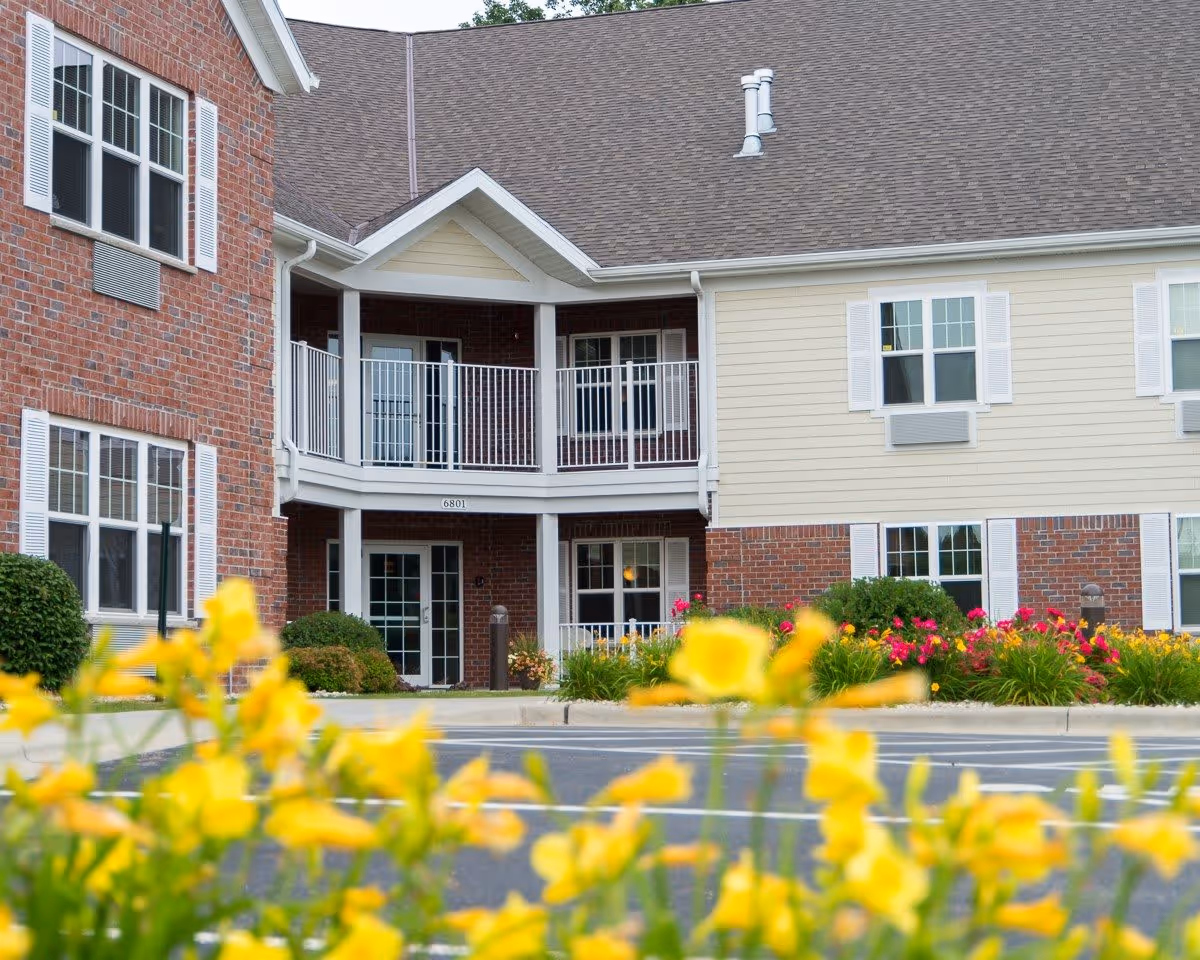 Exterior view of a residential building with brick and beige siding, white window shutters, and a balcony. Yellow flowers and green bushes are in the foreground near a paved area.