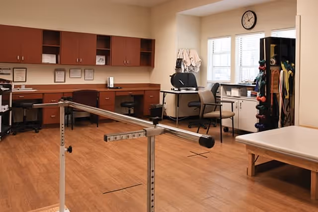 A physical therapy room with wooden flooring, parallel bars for walking exercises, a treatment table, chairs, cabinets, and a rack holding various colored dumbbells. There is a clock on the wall above a window with blinds, and some medical equipment and towels are visible on a counter.