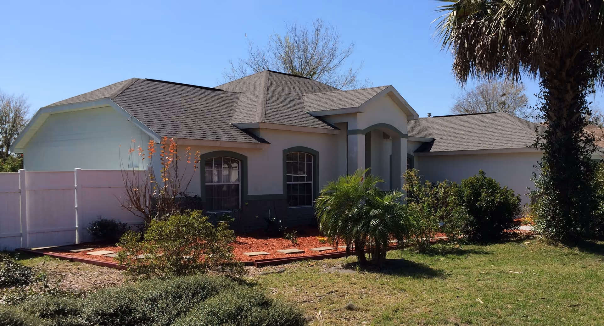 Front exterior of a single-story house with a small landscaped yard, palm tree, and white privacy fence under a clear blue sky.