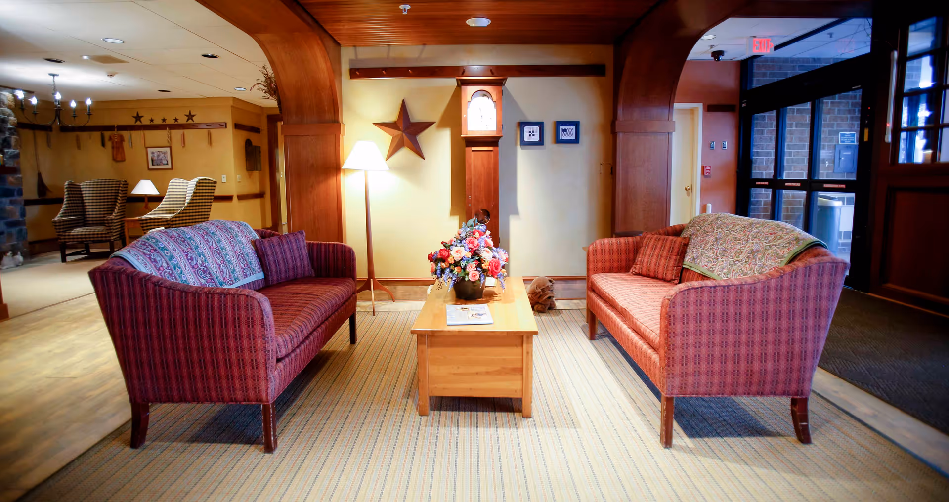 A cozy living room area with two red patterned sofas facing each other across a wooden coffee table with a floral arrangement. The room features wooden arches, a grandfather clock, a floor lamp, and framed pictures on the wall. In the background, there are additional seating areas with armchairs and a stone fireplace, as well as a glass door entrance.