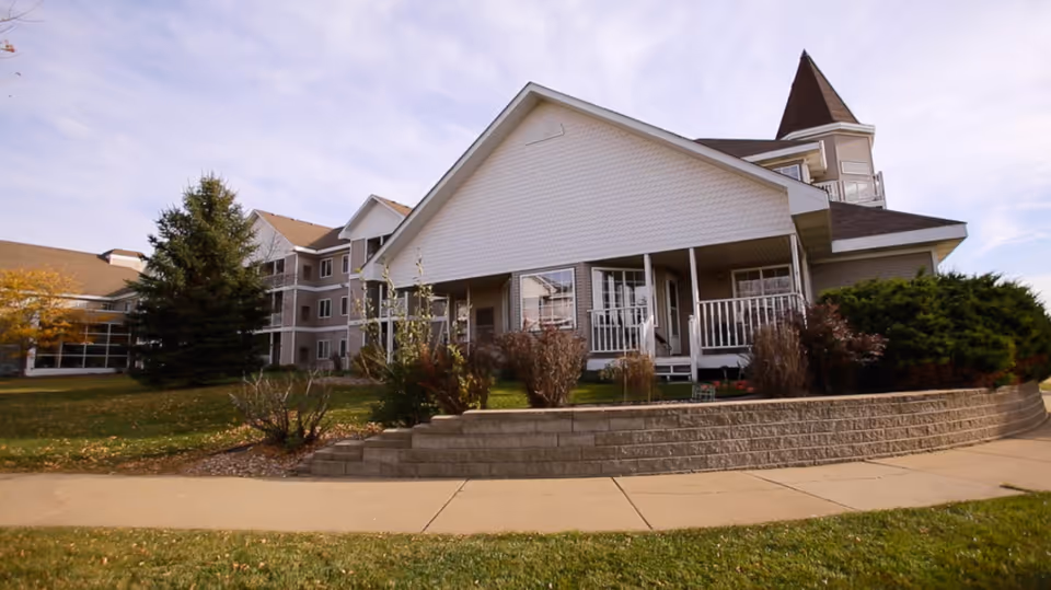 Exterior view of a senior living community building with a white porch, multiple windows, and a pointed tower structure. The building is surrounded by a lawn, bushes, and trees under a partly cloudy sky.