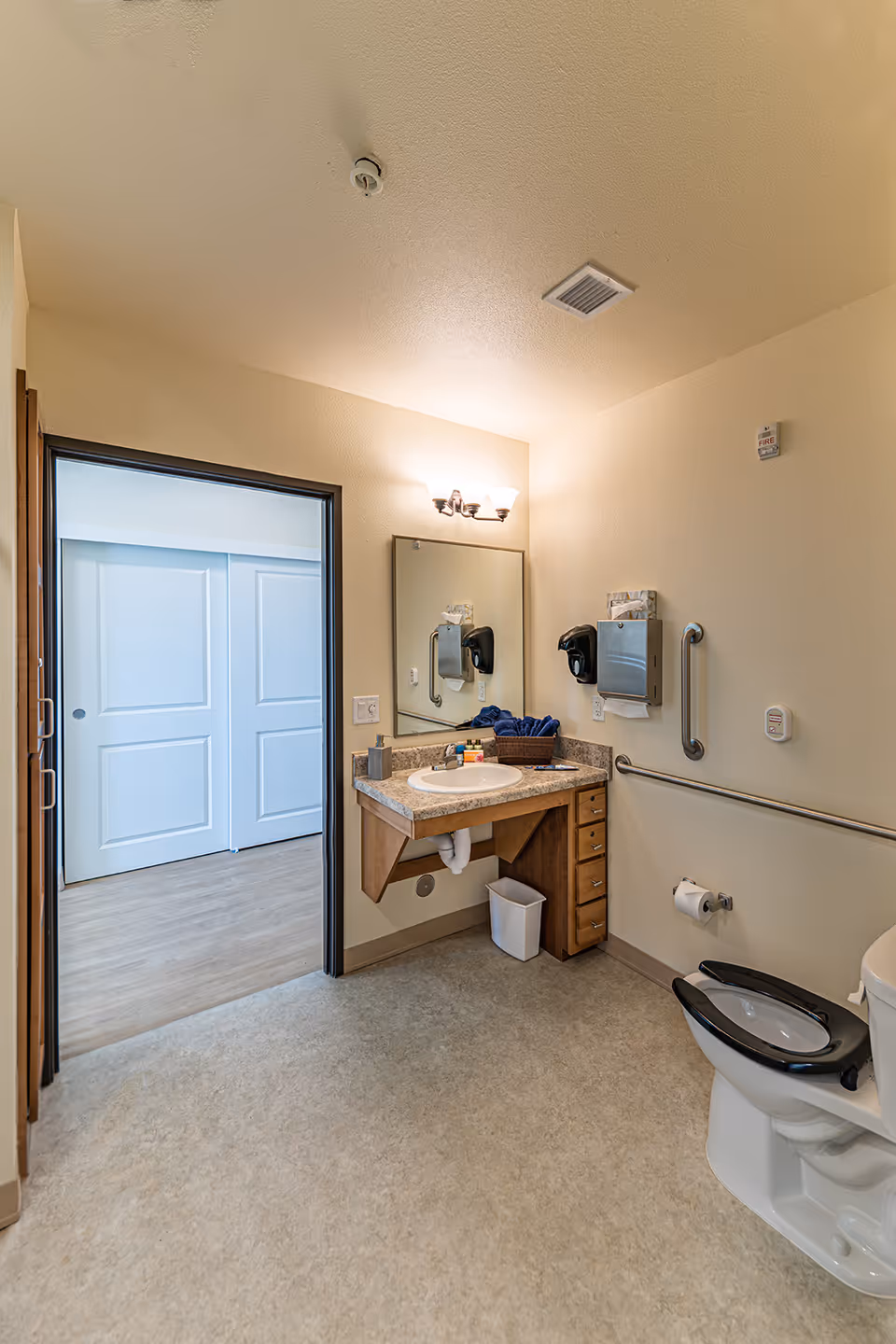 A clean and accessible bathroom with a toilet featuring a raised black seat, a wall-mounted sink with a granite countertop, a large mirror above the sink, and grab bars on the walls. The bathroom has beige walls and flooring, a light fixture above the mirror, and a doorway leading to a room with light wood flooring and white sliding doors.