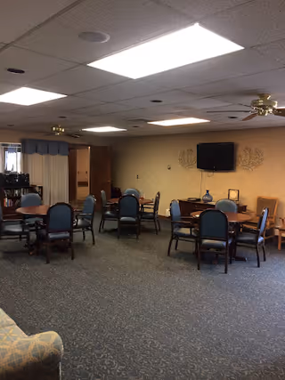A common area in a senior living facility with multiple round tables surrounded by blue cushioned chairs. The room has a carpeted floor, beige walls, ceiling fans, and fluorescent lighting. A flat-screen TV is mounted on the wall, and there is a small side table with decorative items beneath it.