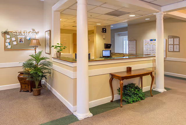 Reception desk area in a senior living facility lobby with white columns, a console table, potted plants, and a bulletin board.