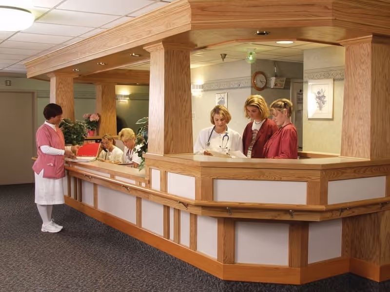 A nursing facility reception area with a wooden front desk. Four healthcare workers, including nurses and staff, are engaged in reviewing documents behind the desk. One nurse in a pink uniform stands on the visitor side of the desk. The area is well-lit with ceiling lights and decorated with plants and framed artwork on the walls.