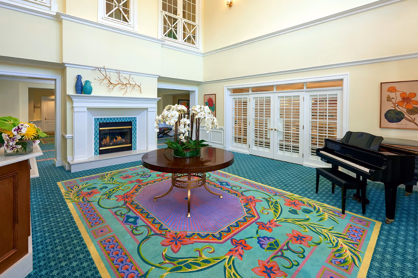 A brightly lit living room area with a colorful floral patterned carpet, a round wooden table with white orchids in the center, a black grand piano on the right, a fireplace with blue tiles and decorative vases on the mantel, and white French doors with shutters. The walls are painted light yellow and there is a framed floral artwork on the right wall.
