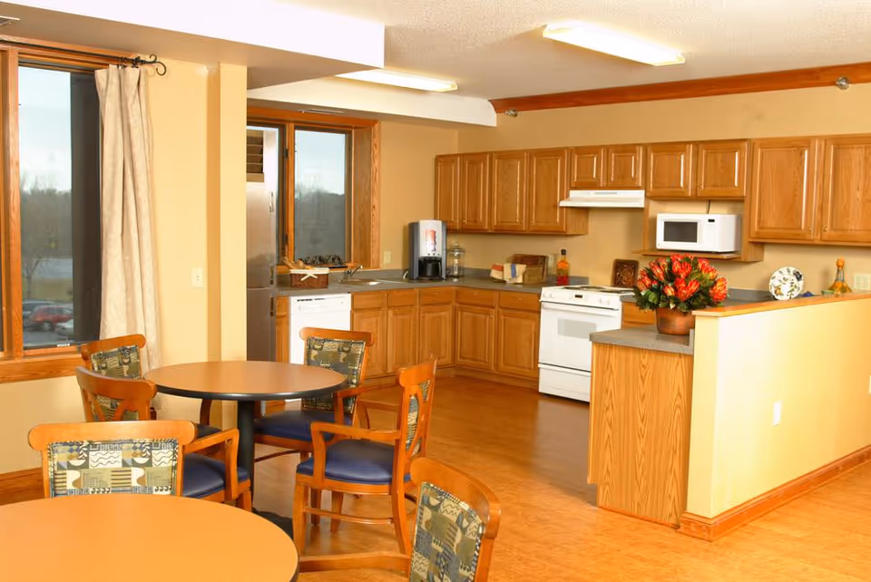 A bright kitchen and dining area with wooden cabinets, a white stove, microwave, and dishwasher. There are two round tables with wooden chairs that have patterned cushions. A large window with beige curtains lets in natural light, and a vase with red flowers sits on the kitchen counter.