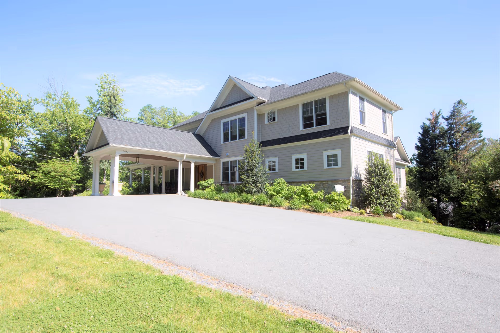 A two-story residential building with a covered driveway entrance, surrounded by greenery and trees under a clear blue sky.