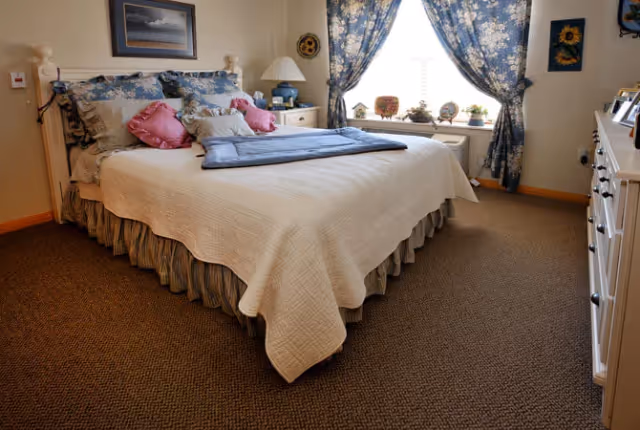 A cozy bedroom with a large bed covered in a white quilt and multiple pillows in shades of pink and blue. The room has a window with floral blue curtains, a nightstand with a lamp, and a dresser with framed photos. The carpet is brown and the walls are light-colored.