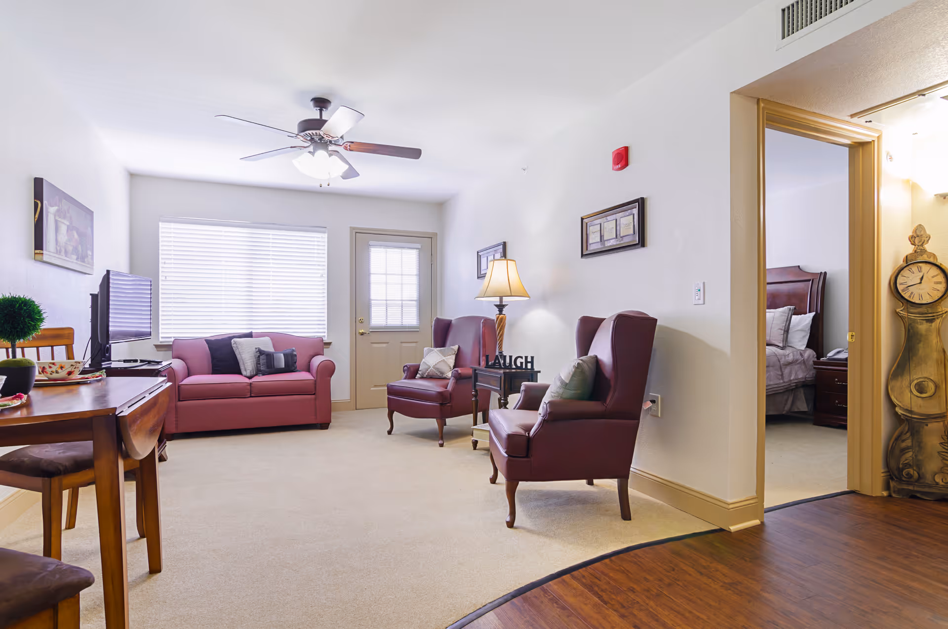 A bright living room in an assisted living facility featuring a ceiling fan with lights, a pink loveseat with pillows, two burgundy armchairs with pillows, a wooden side table with a lamp and decorative items, a wooden dining table with chairs, a TV on a stand, and an open doorway leading to a bedroom with a bed and nightstand. The room has beige carpet and white walls with framed artwork.