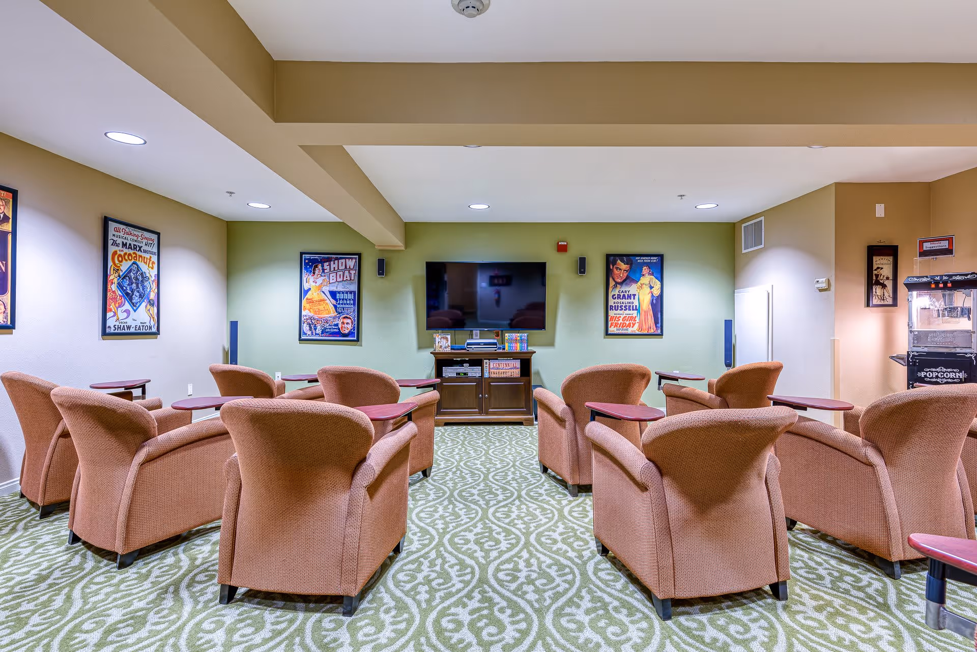A cozy entertainment room with eight cushioned armchairs arranged in two rows facing a flat-screen TV mounted on a light green wall. The room has vintage movie posters on the walls, a patterned green carpet, and a popcorn machine in the corner.