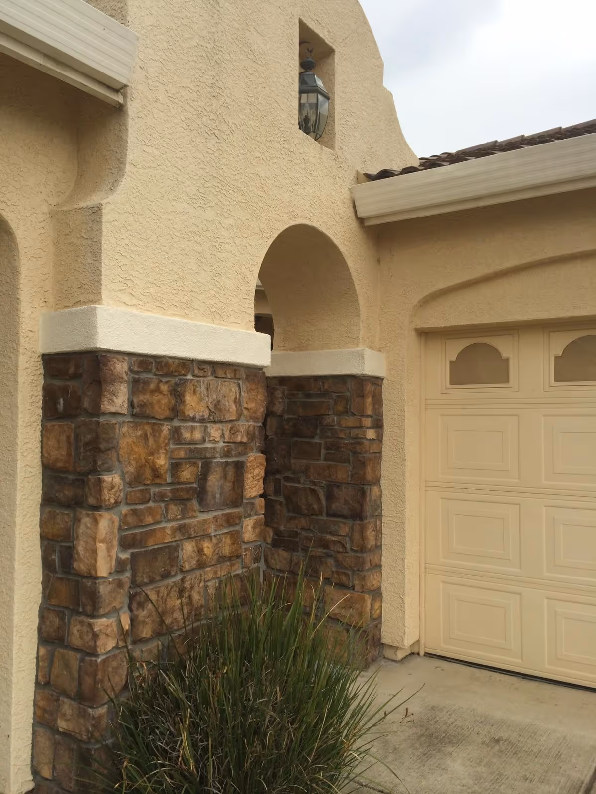 Exterior view of a building corner showing a beige stucco wall with stone accents, an arched architectural detail, a lantern-style light fixture, a garage door, and some green shrubbery at the base.