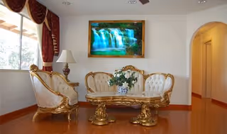 A living room with ornate gold and white upholstered furniture including a sofa and two chairs, a matching coffee table with a floral arrangement, a table lamp on a side table, a large window with red curtains, and a framed picture of a waterfall on the wall.