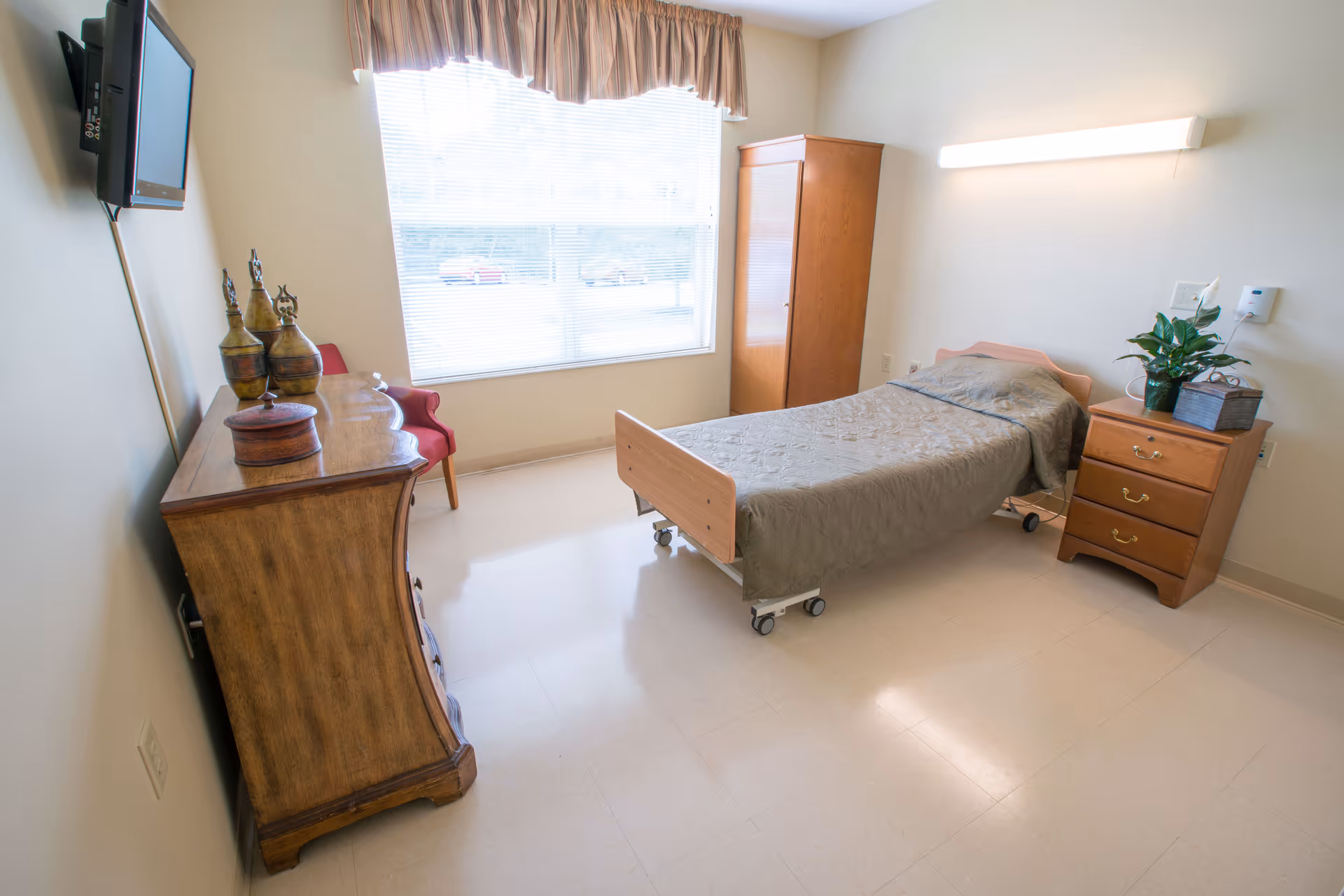 A simple, clean bedroom in a senior living facility with a single bed on wheels covered with a gray quilt. The room has a wooden dresser with decorative items, a wooden wardrobe, a small bedside table with a plant, a red chair near the window, and a wall-mounted TV. The window has blinds and a valance, and the floor is light-colored tile.
