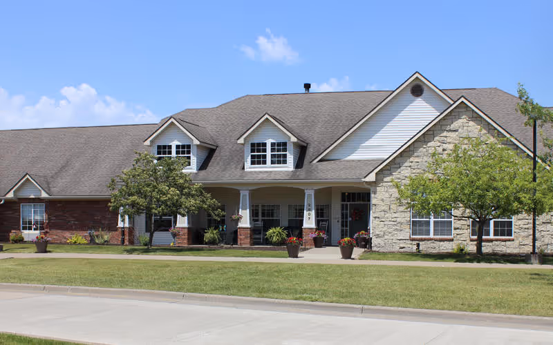 Front exterior view of a single-story senior living facility building with a combination of brick and stone facade, multiple windows, a covered entrance with columns, potted plants, and a well-maintained lawn under a blue sky with some clouds.