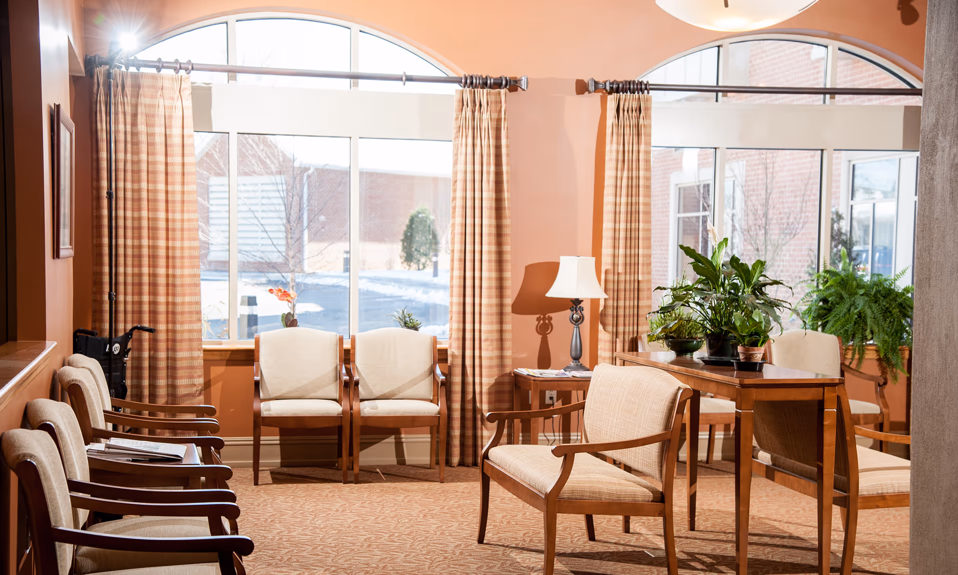 Sunny seating area in a senior living facility with upholstered chairs, a wooden table, potted plants and large arched windows with curtains.