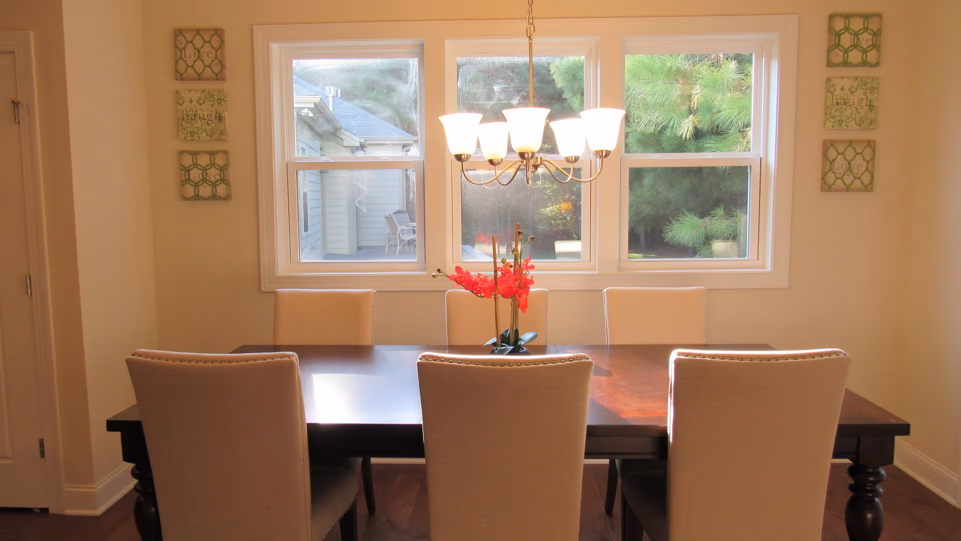 A dining room with a dark wooden table surrounded by six beige upholstered chairs. A chandelier with six lit lamps hangs above the table. A vase with red flowers is placed in the center of the table. Three large windows behind the table show an outdoor view with greenery and part of a building. On each side of the windows, there are three decorative wall plaques with the words 'Live', 'Laugh', and 'Love'.