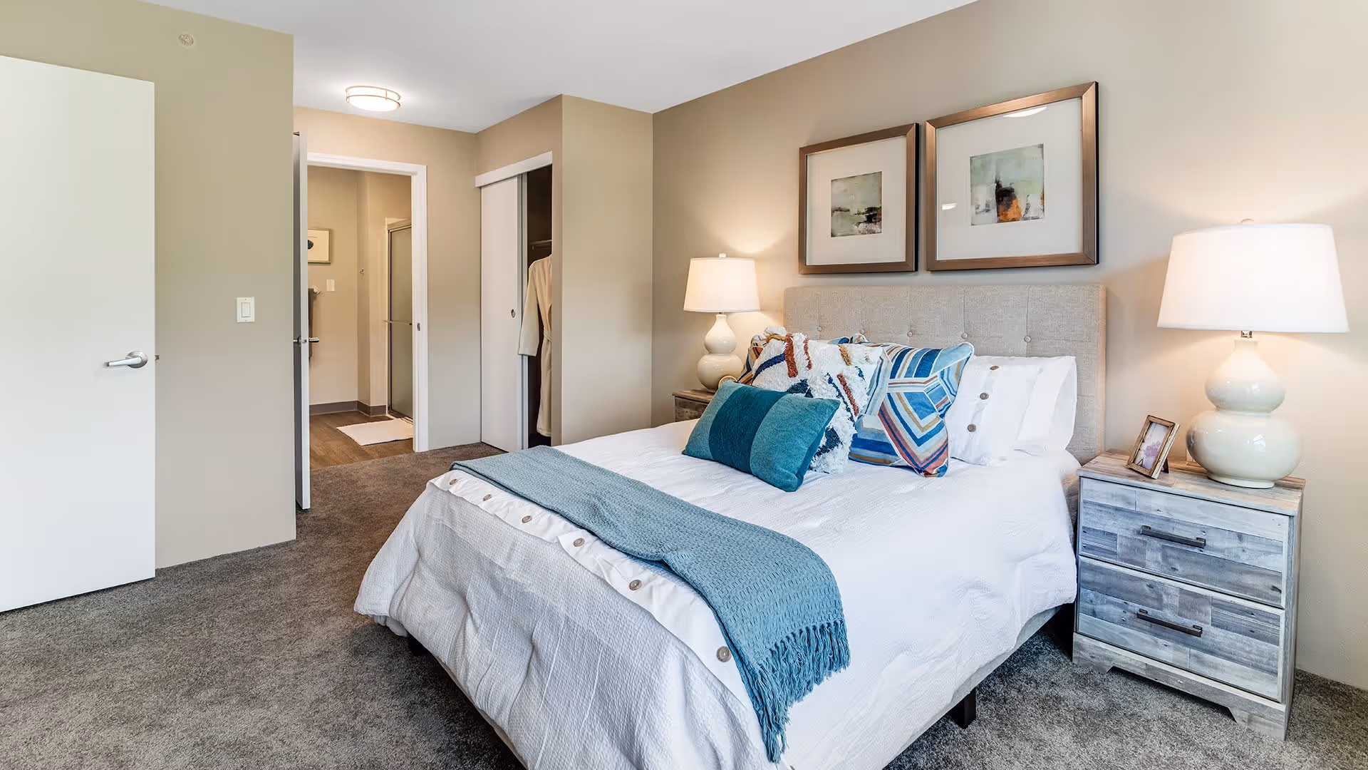 A neatly made bed with white bedding and multiple decorative pillows in shades of blue and patterned designs. There is a blue throw blanket at the foot of the bed. On either side of the bed are wooden nightstands with matching lamps. Two framed abstract artworks hang above the bed. The room has beige walls and carpeted flooring. An open door and a closet with sliding doors are visible, leading to another room.