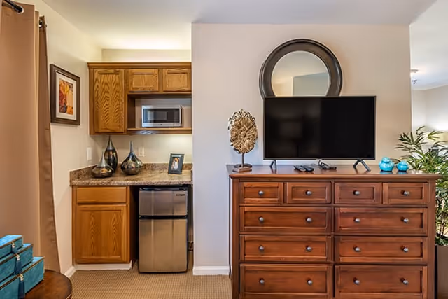 Interior room showing a kitchenette with a microwave and mini-fridge beside a wooden dresser topped with a flat-screen TV and decorative items.