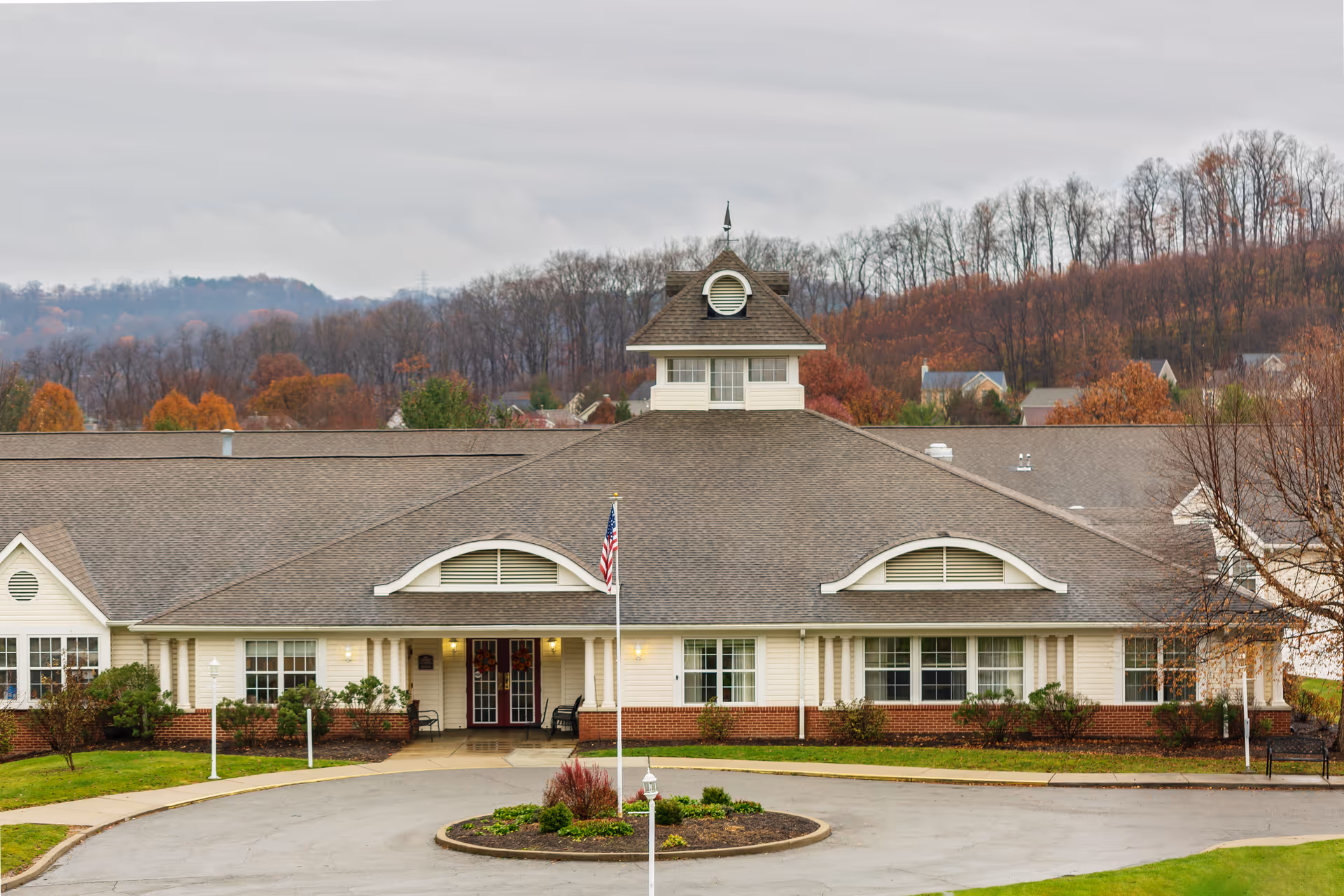 Front exterior view of Celebration Villa of Lakemont Farms, a single-story building with a peaked roof and a cupola on top. The entrance has double doors with wreaths, flanked by windows and columns. There is a circular driveway with a flagpole displaying the American flag in front, and trees with autumn foliage in the background.