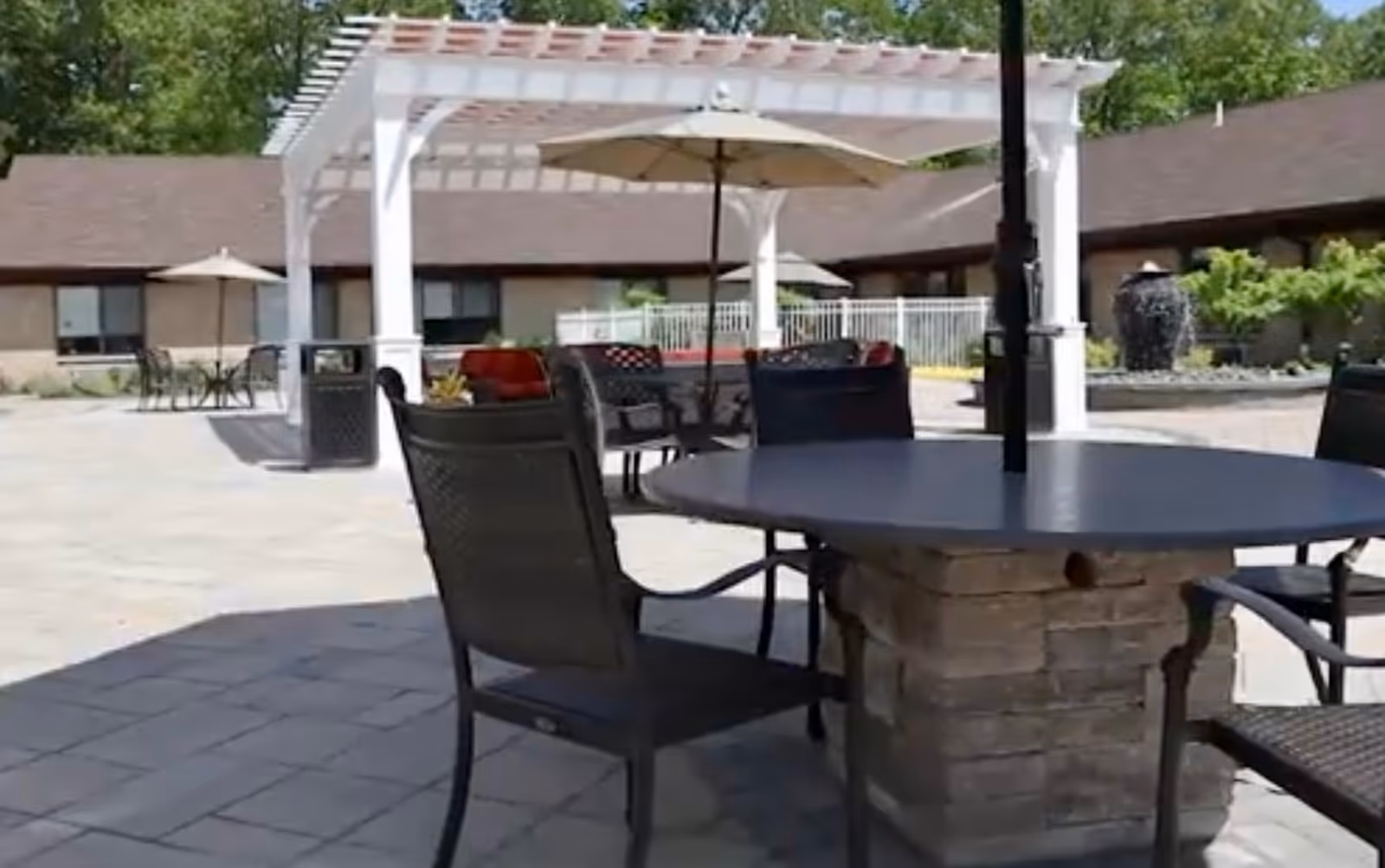 Outdoor patio area with round tables and chairs, some tables have umbrellas. There is a white pergola structure with seating underneath and a building with windows in the background surrounded by greenery.