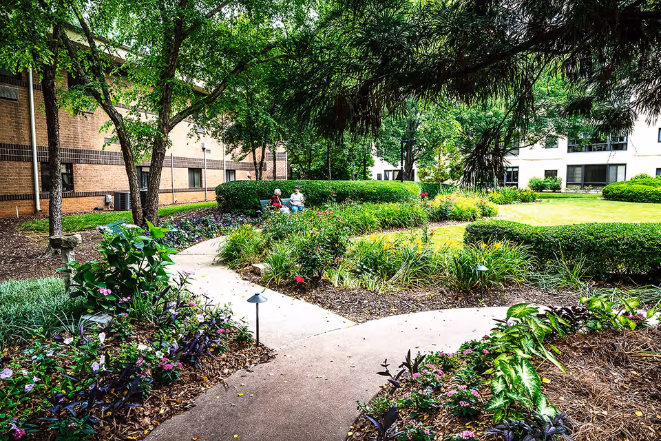 A landscaped outdoor garden area at Montclair Assisted Living with a paved walkway, green bushes, flowering plants, and trees. Two elderly people are sitting on a bench in the background near the building.